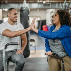 Happy plump African American female trainee and muscular smiling coach clapping each others hands while sitting on stationary bicycles in modern gym