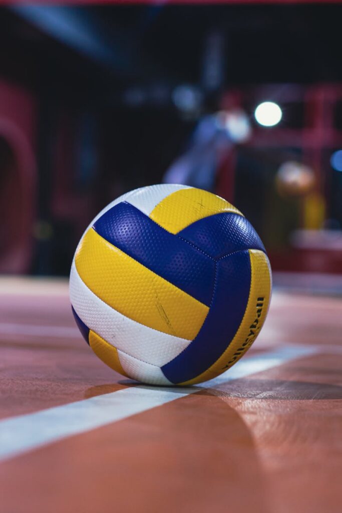 Yellow and white volleyball resting on an indoor volleyball court with bright lights.