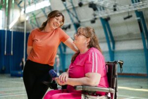 Two women, one in a wheelchair, smiling and interacting in a gym setting.