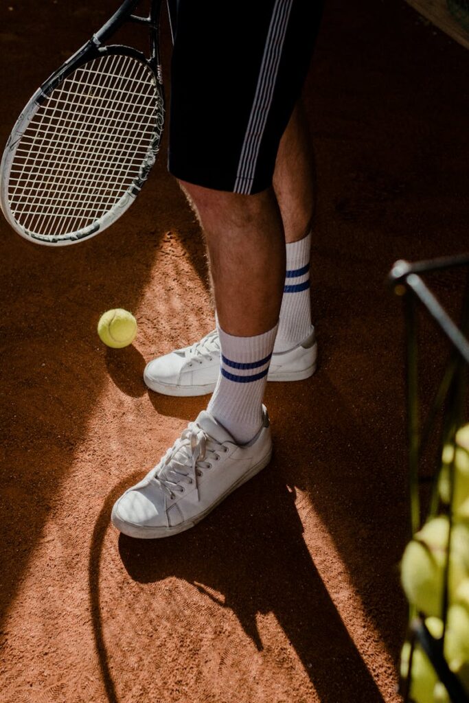 A tennis player stands on a clay court, showing sneakers, racket, and tennis balls in sunlight.