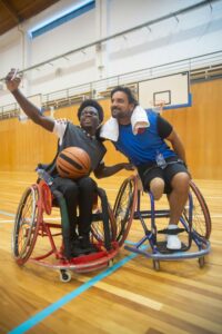 Two wheelchair basketball players smiling and taking a selfie on the court.